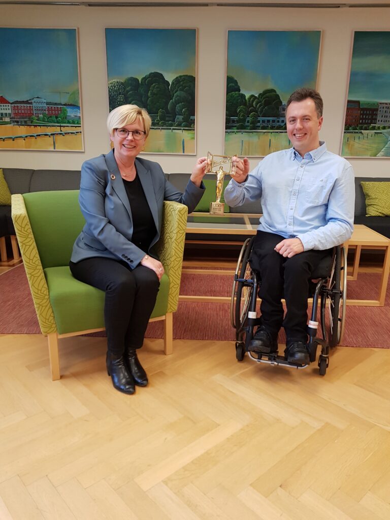 Bild på kommunalrådet Ann-Marie Nilsson och Funktionsrätts Johan Steirud. Sitter ner och håller båda i priset mellan sig.